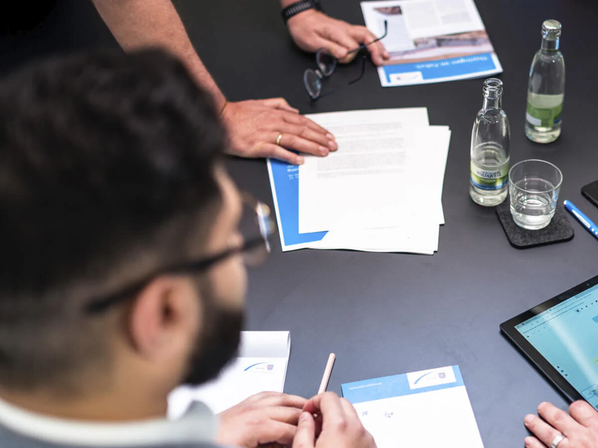 Weiterbildung Bildbeschreibung / description: Menschen beraten sich an einem Tisch mit einem Tablet und Unterlagen.